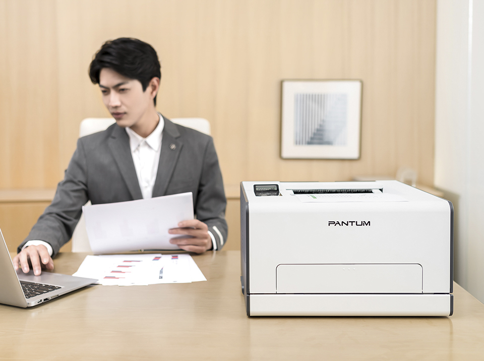Máy in Pantum CP2100DW trên bàn làm việc cùng tài liệu đã in và người đang làm việc bên máy tính xách tay